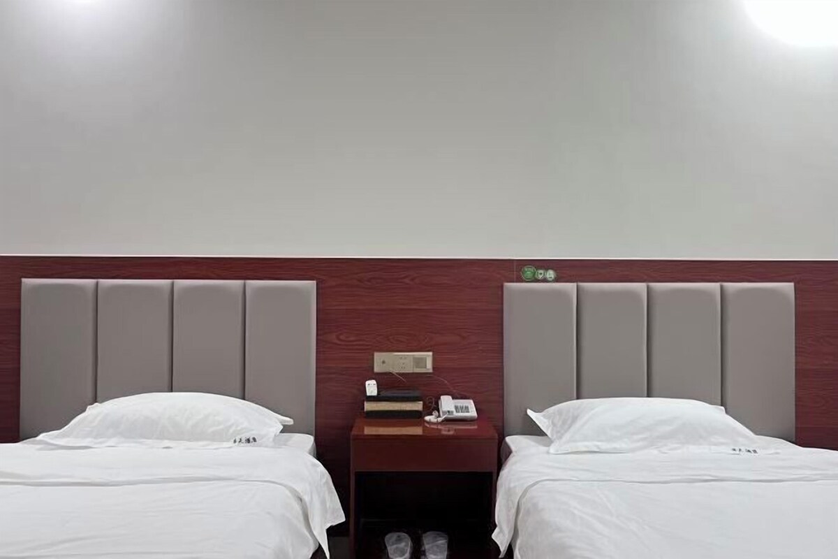 A hotel room with two beds, a wooden headboard, a nightstand with a phone and books, and a small shelf with shoes.