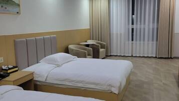 A hotel room with two beds, a headboard, a small table with a chair, and a window with curtains.