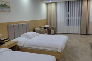 A hotel room with two beds, a headboard, a small table with a chair, and a window with curtains.