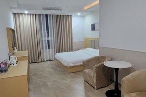 A hotel room with a bed, a desk with toiletries, a round table, and a large window with curtains.