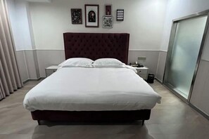 A neatly made bed with white linens, a dark red headboard, and a small bedside table with a lamp.