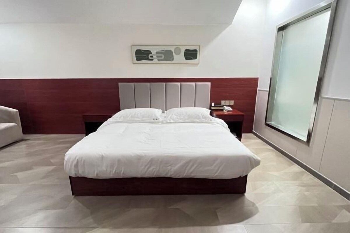 A modern hotel room with a large bed, a wooden headboard, a nightstand, and a large window providing a view.