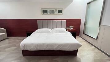 A modern hotel room with a large bed, a wooden headboard, a nightstand, and a large window providing a view.