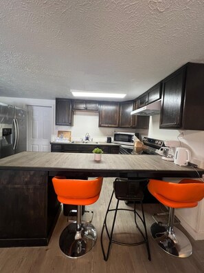 Una cocina moderna con armarios de madera oscura, un refrigerador de acero inoxidable, un microondas y una estufa. Hay dos taburetes naranja y una pequeña planta en maceta en la isla.