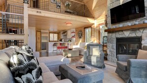 A spacious living area with a stone fireplace, a kitchen in the background, and a balcony with a railing. - Baycliff Kanata Tremblant (257) (Amherst)