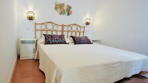 Una cama con cubrecama blanco y dos almohadas estampadas, cabecera de madera, dos mesitas de noche con lámparas y una decoración en la pared sobre la cama.