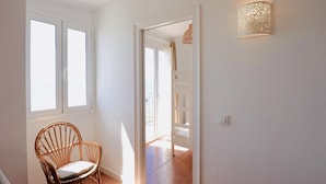 Un pasillo luminoso con una silla de mimbre, una puerta que da a un balcón y una lámpara fijada a la pared.