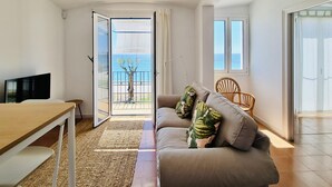 Una sala de estar moderna con un sofá, un televisor, una mesa de madera y vistas al mar a través de una puerta abierta.