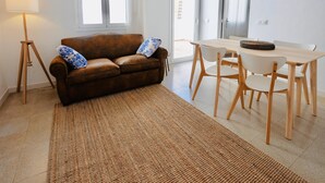 A living room with a brown leather sofa, a wooden dining table with white chairs, a woven floor rug, and a lamp. - Mundet 4 Dalt Terrassa. CostaBravaSi - At the sea (Sant Antoni de Calonge)