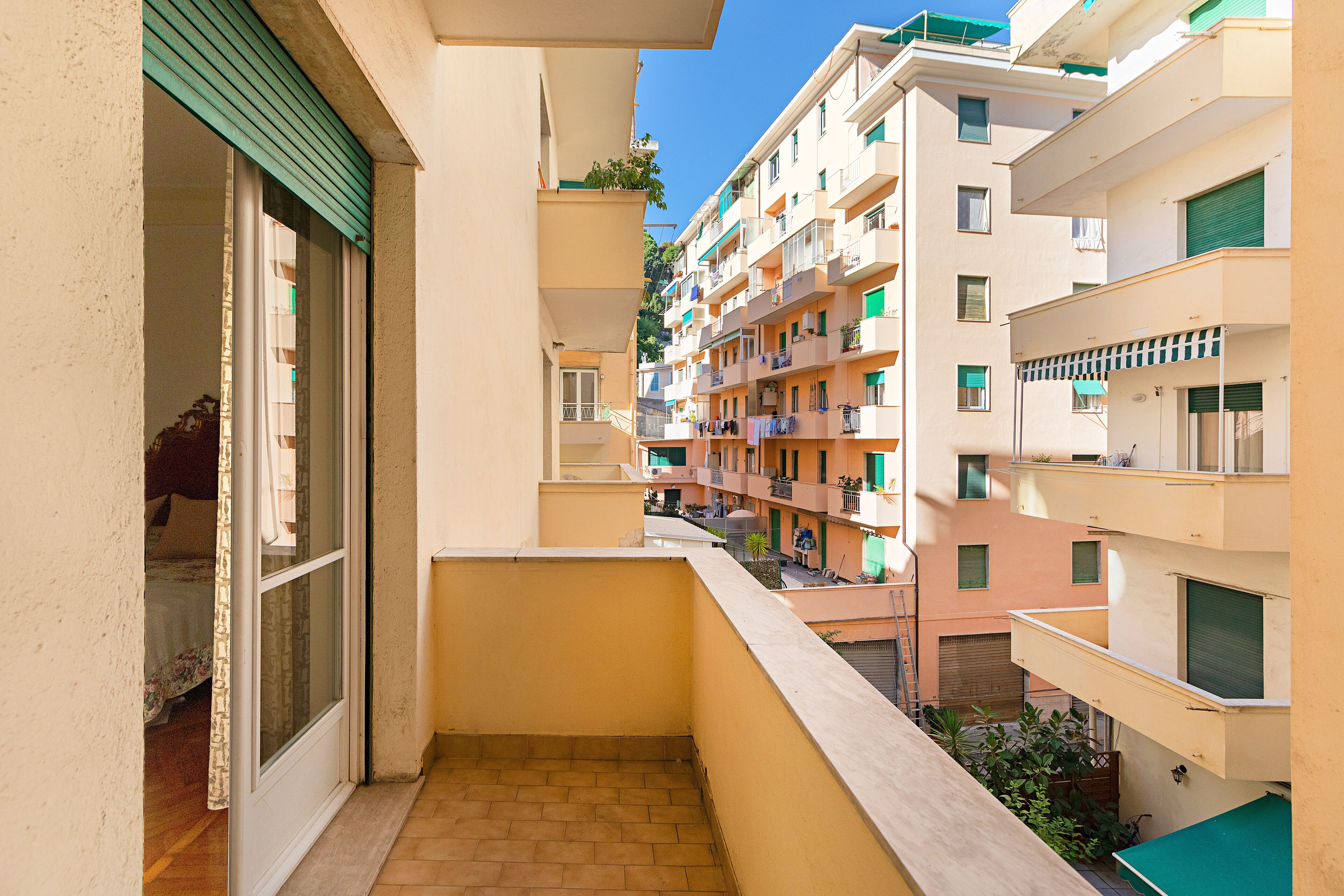 A narrow balcony with a view of adjacent buildings, a door leading to a room, and a small outdoor area with a plant.