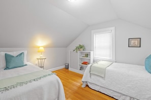 Un dormitorio con dos camas, una mesita de noche con lámpara, una ventana con persianas, una estantería y un cuadro en la pared.