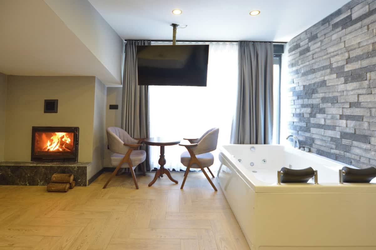 A modern living room with a fireplace, a flat-screen TV, a stone wall, a large window with curtains, and a jacuzzi tub.