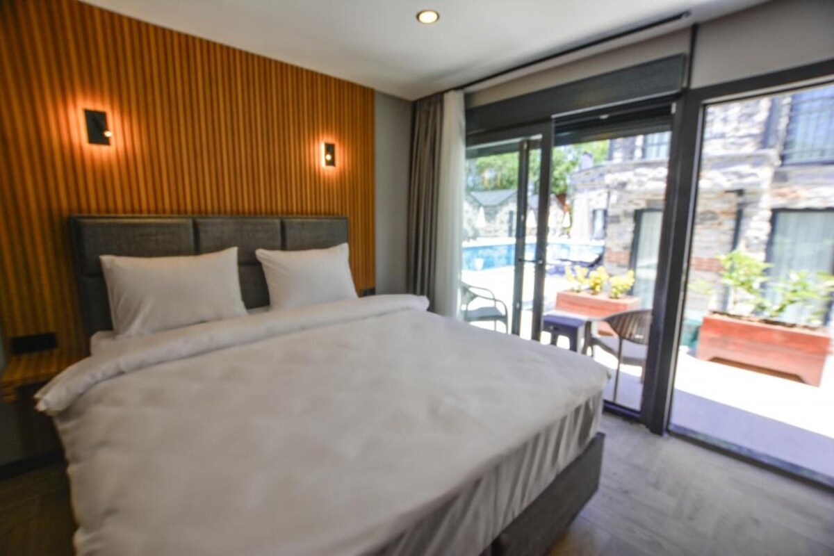 A modern hotel room with a large bed, wooden paneling on the walls, and a sliding glass door leading to an outdoor area with tables and chairs.