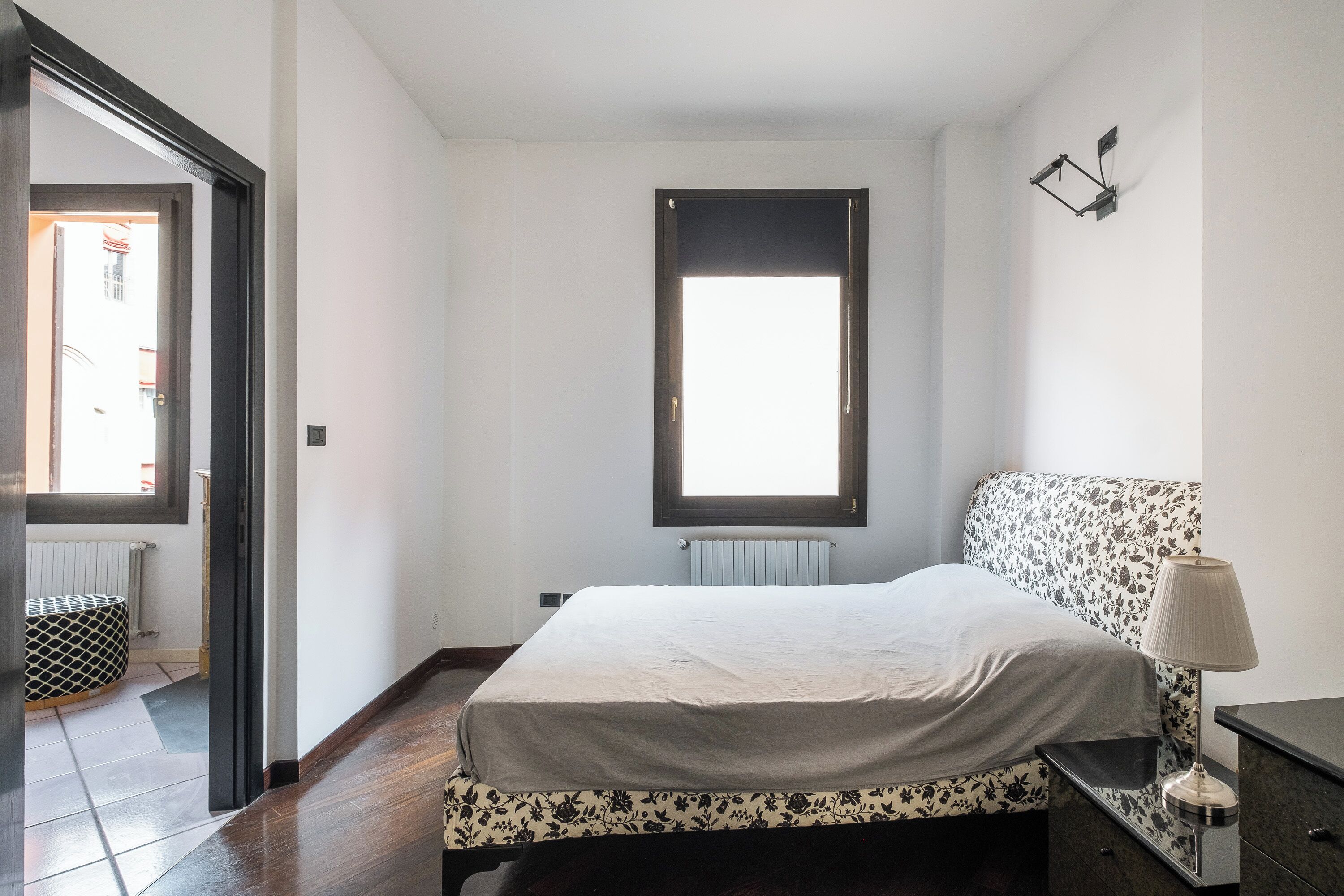 Une chambre à coucher avec un lit, une table de chevet, une lampe, une fenêtre et un radiateur.