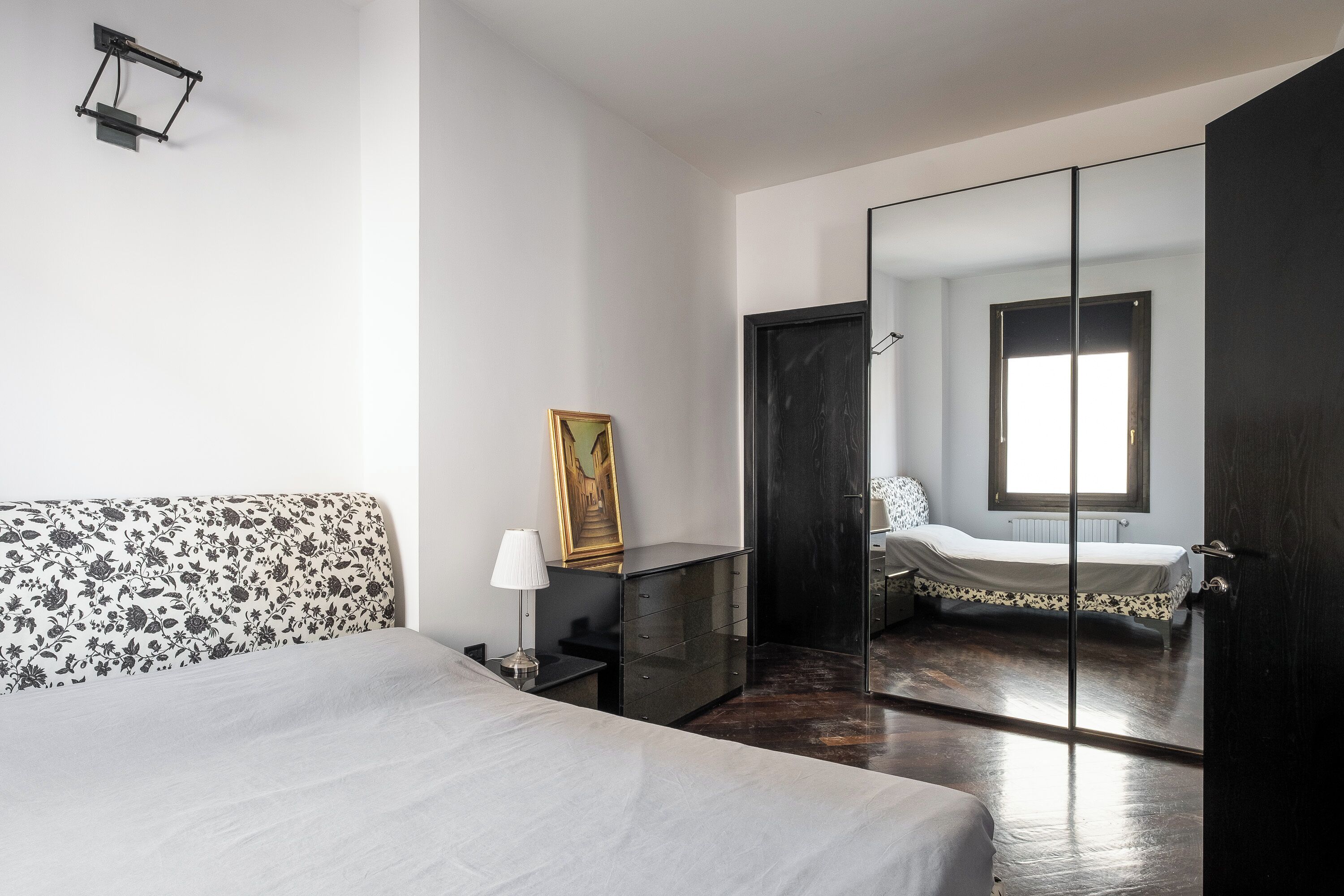 A bedroom with a bed, a bedside table, a lamp, a dresser, a mirror, and a door leading to another room.