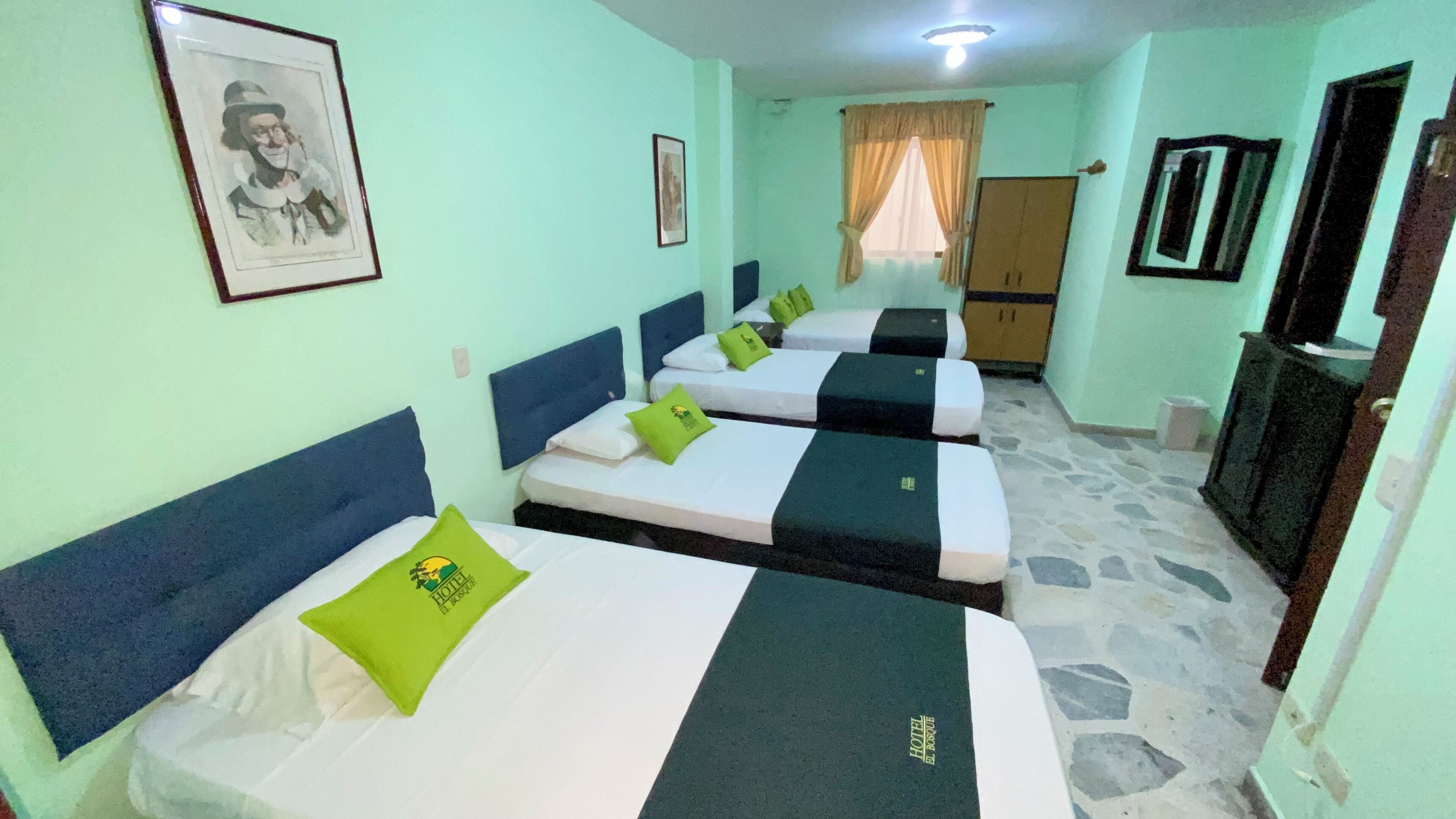 A hotel room with four single beds, each with a pillow and a headboard. There is a wardrobe, a television, and a framed picture on the wall.