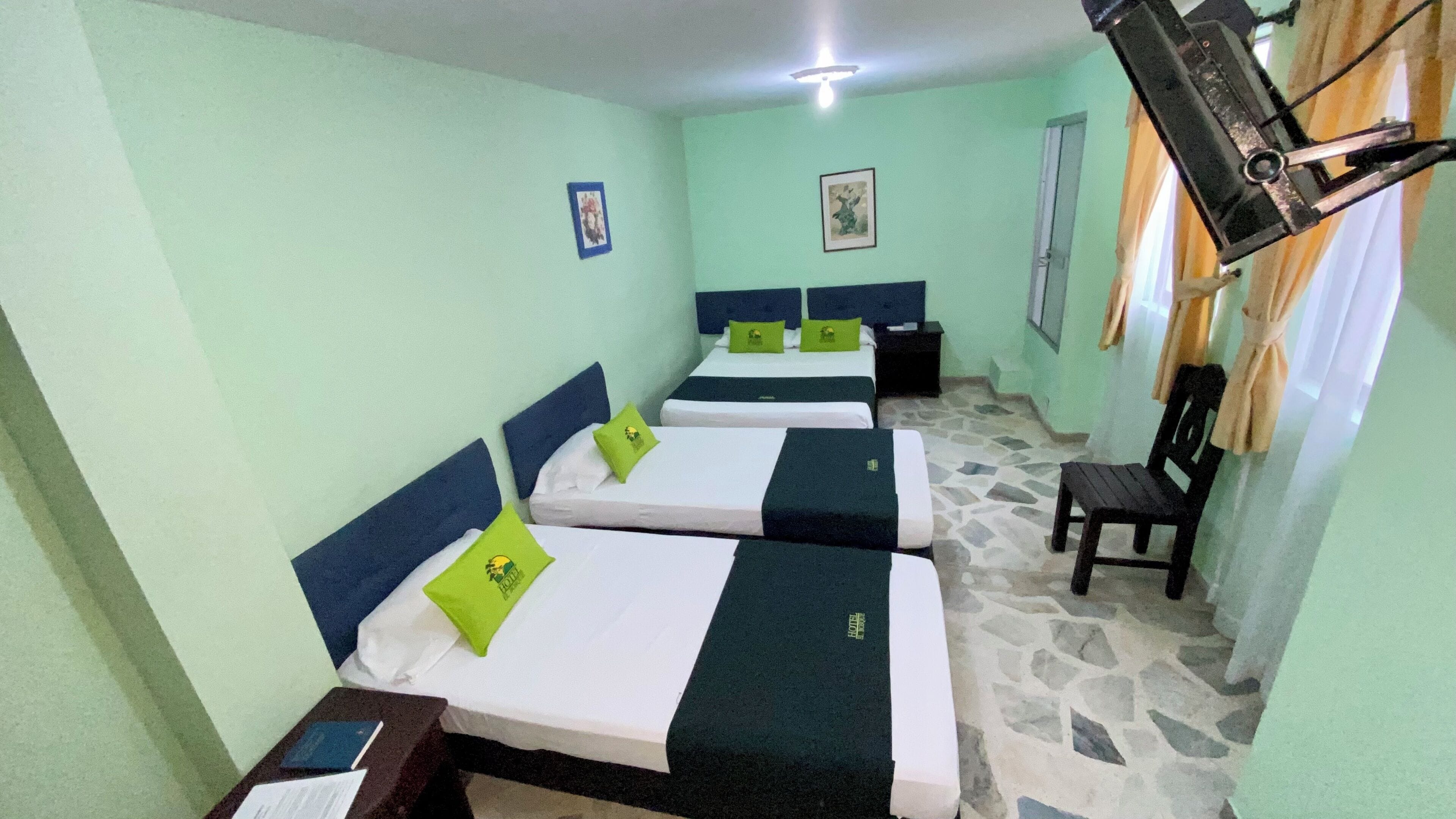 A room with three beds, each with a green pillow and a headboard. There is a wooden chair and a small table with a lamp. The walls are painted light green, and there is a window with curtains.