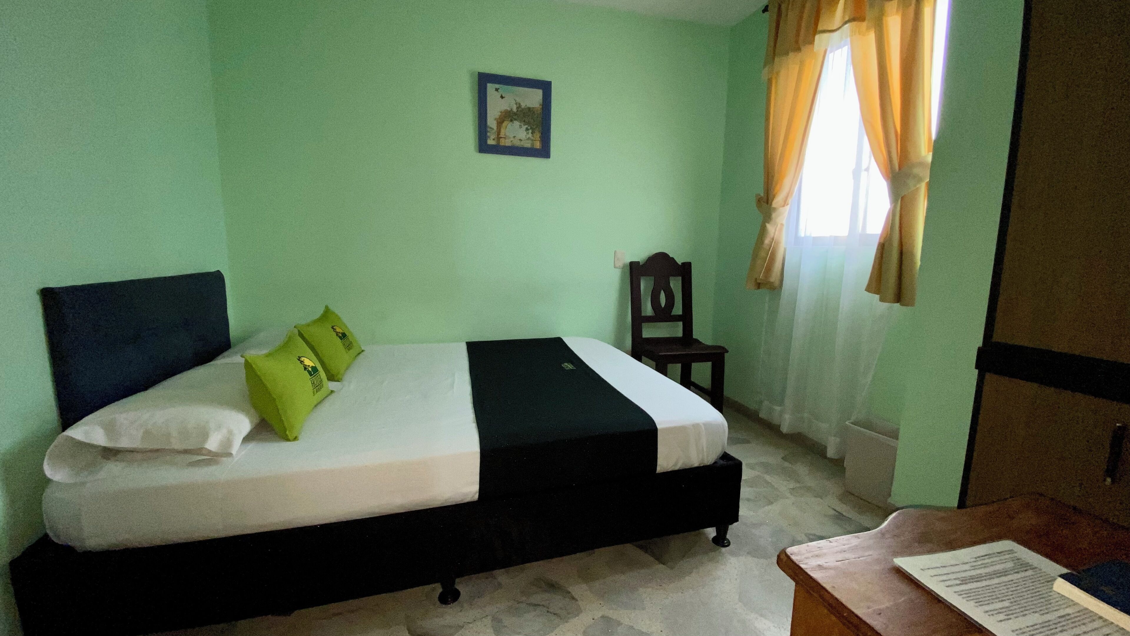 A room with a bed, a chair, a table with a book, a picture on the wall, and a window with curtains.