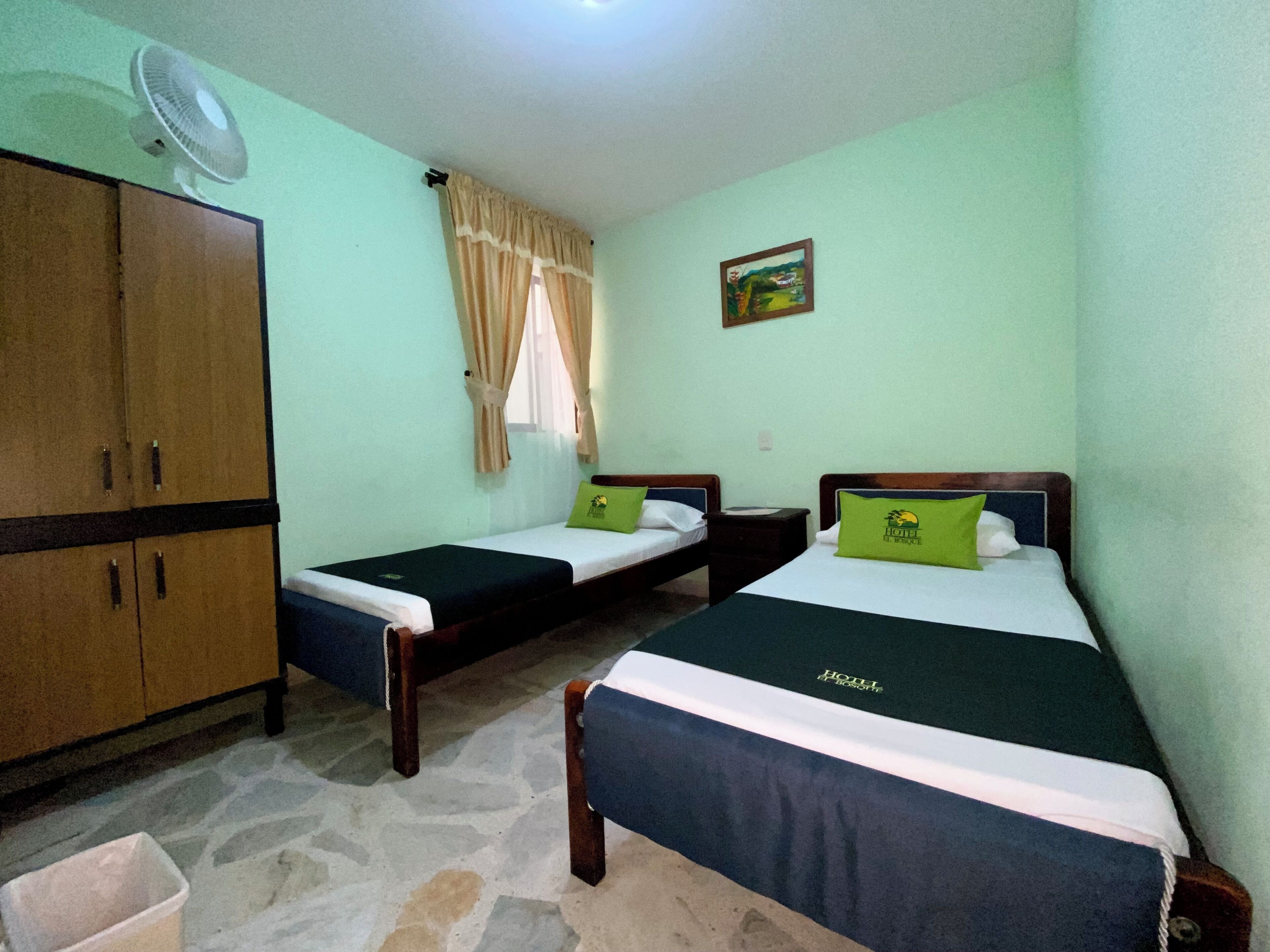 Une chambre d’hôtel avec deux lits simples, une armoire, un ventilateur et une fenêtre avec des rideaux.