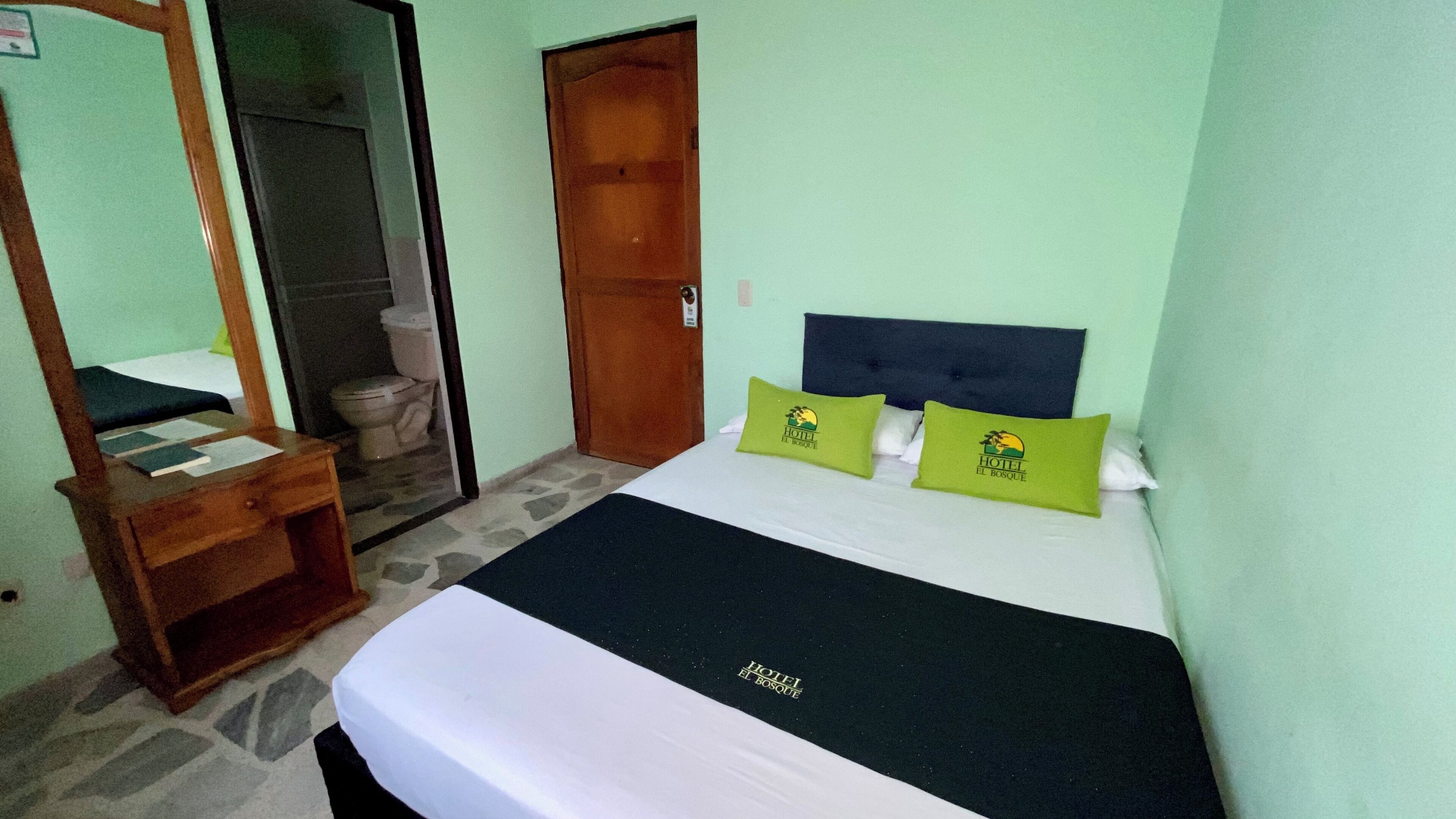 A hotel room with a bed, a wooden nightstand, a mirror, and a bathroom visible through an open door.