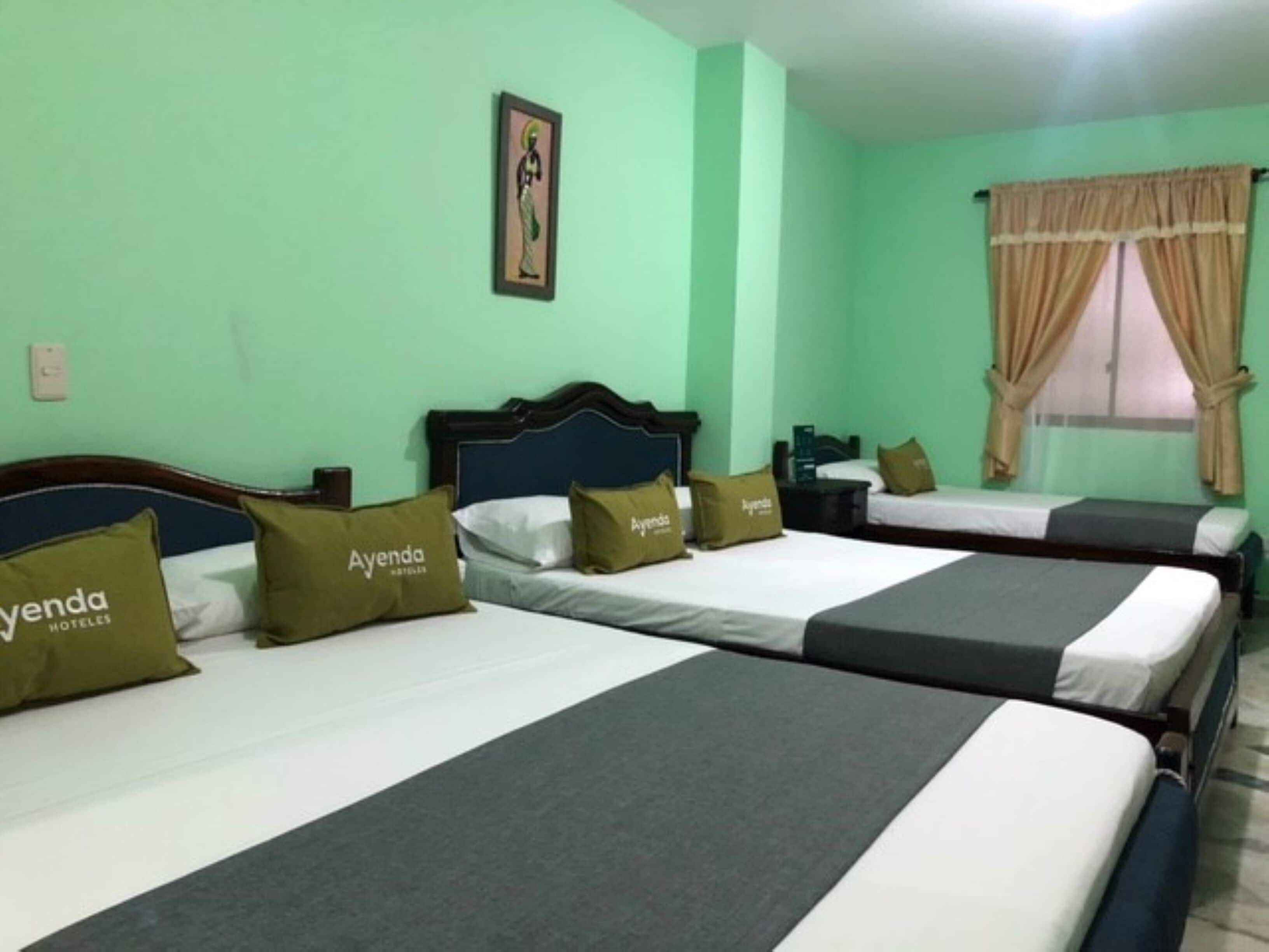 A hotel room with three beds, each with a grey and white bedspread and pillows labeled "Ayenda Hoteles".