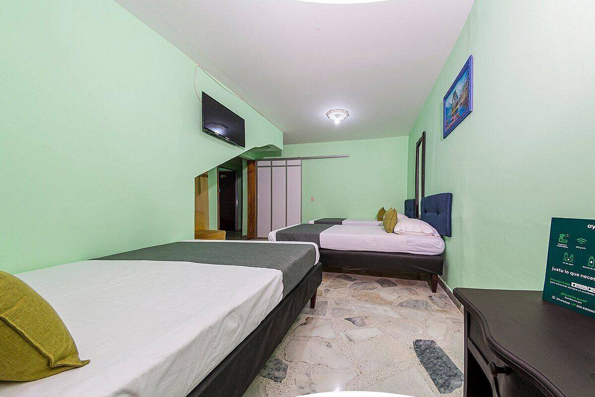 A small hotel room with two single beds, a flat-screen TV mounted on the wall, a small desk with a chair, and a tiled floor.