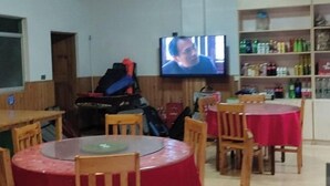 Une pièce avec un téléviseur fixé au mur, des tables en bois recouvertes de nappes rouges et des étagères remplies de divers objets.