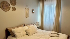 A neatly made bed with pillows and folded towels, a wall-mounted macrame piece, and a sunburst decoration.