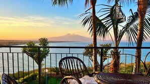 A balcony with wicker chairs and a table, overlooking a body of water and a distant mountain.