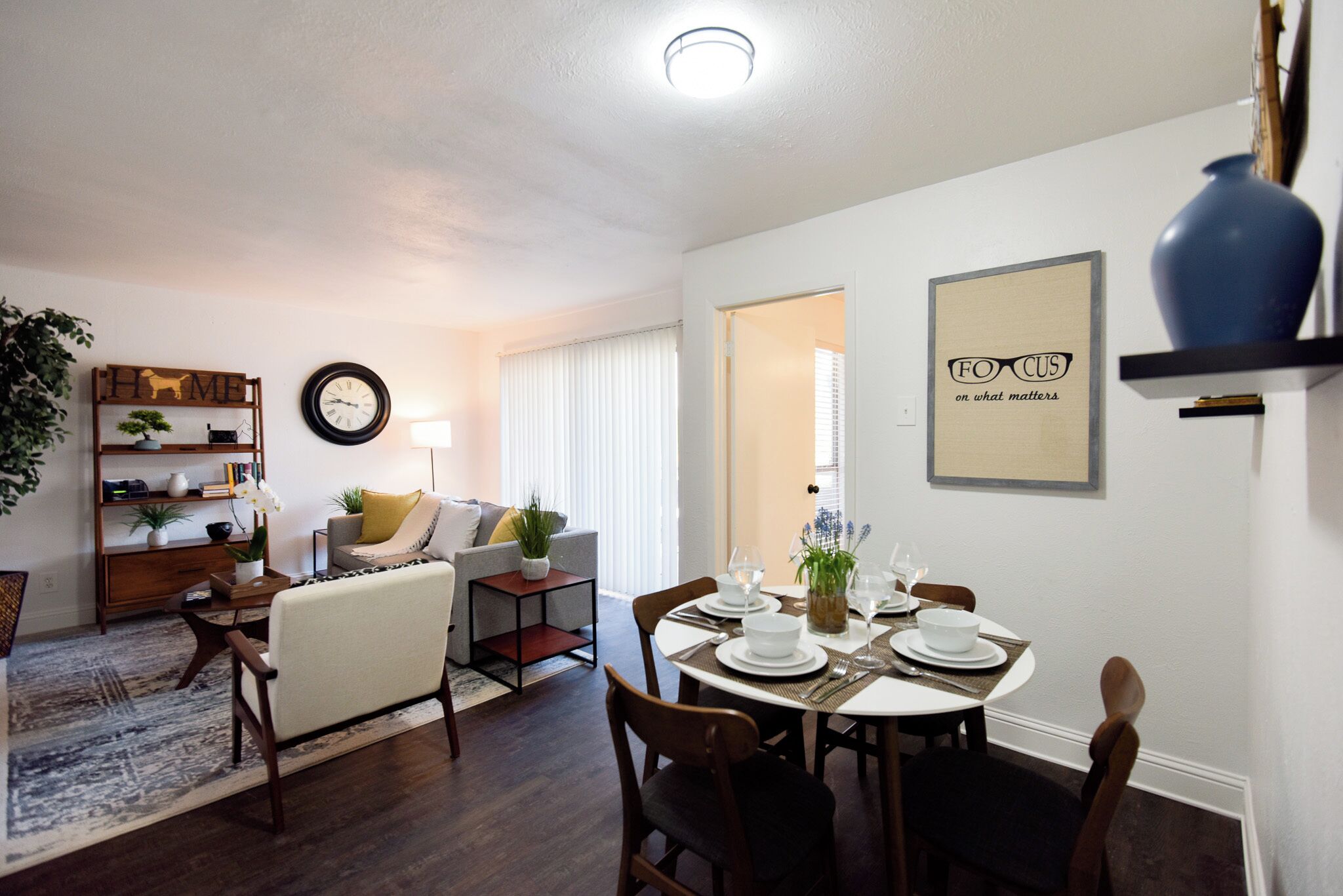 A dining area with a round table set for four, surrounded by chairs. There is a sofa and a small side table with a lamp in the background.