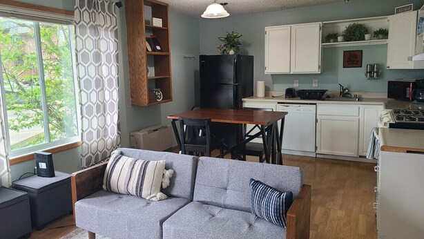 Un espacio habitable compacto con un sofá gris, una mesa de comedor y una cocina con armarios blancos y un refrigerador negro.