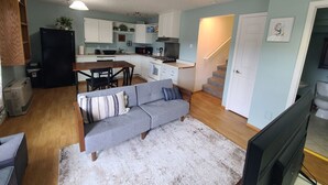 Una sala de estar con un sofá gris, un tapete blanco y piso de madera. Se ve una cocina al fondo con armarios blancos y un refrigerador negro.