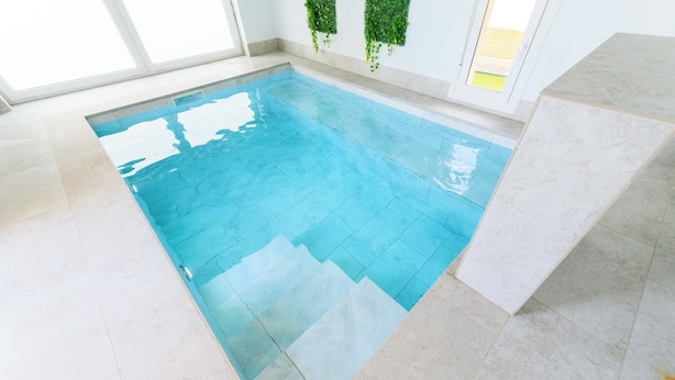 Une piscine intérieure à l’eau d’un bleu limpide, dotée d  ’un sol carrelé et offrant une vue sur le ciel grâce à de grandes fenêtres.