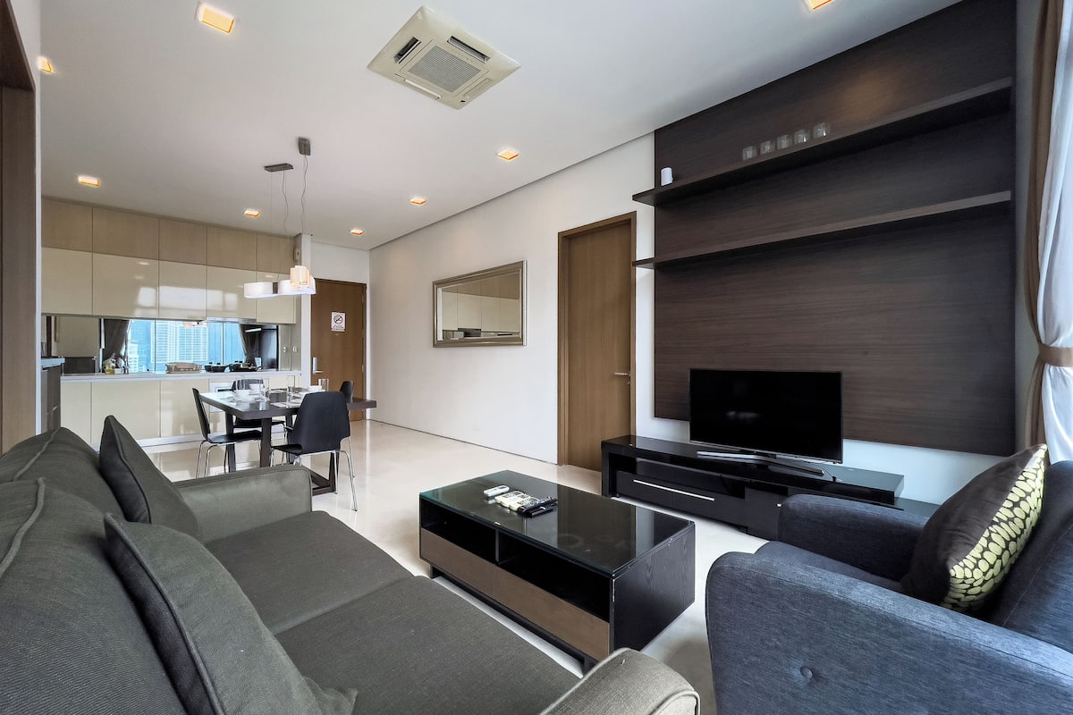 Un salon moderne avec un canapé, une table basse, un téléviseur et un coin repas, la cuisine étant visible à l’arrière-plan.