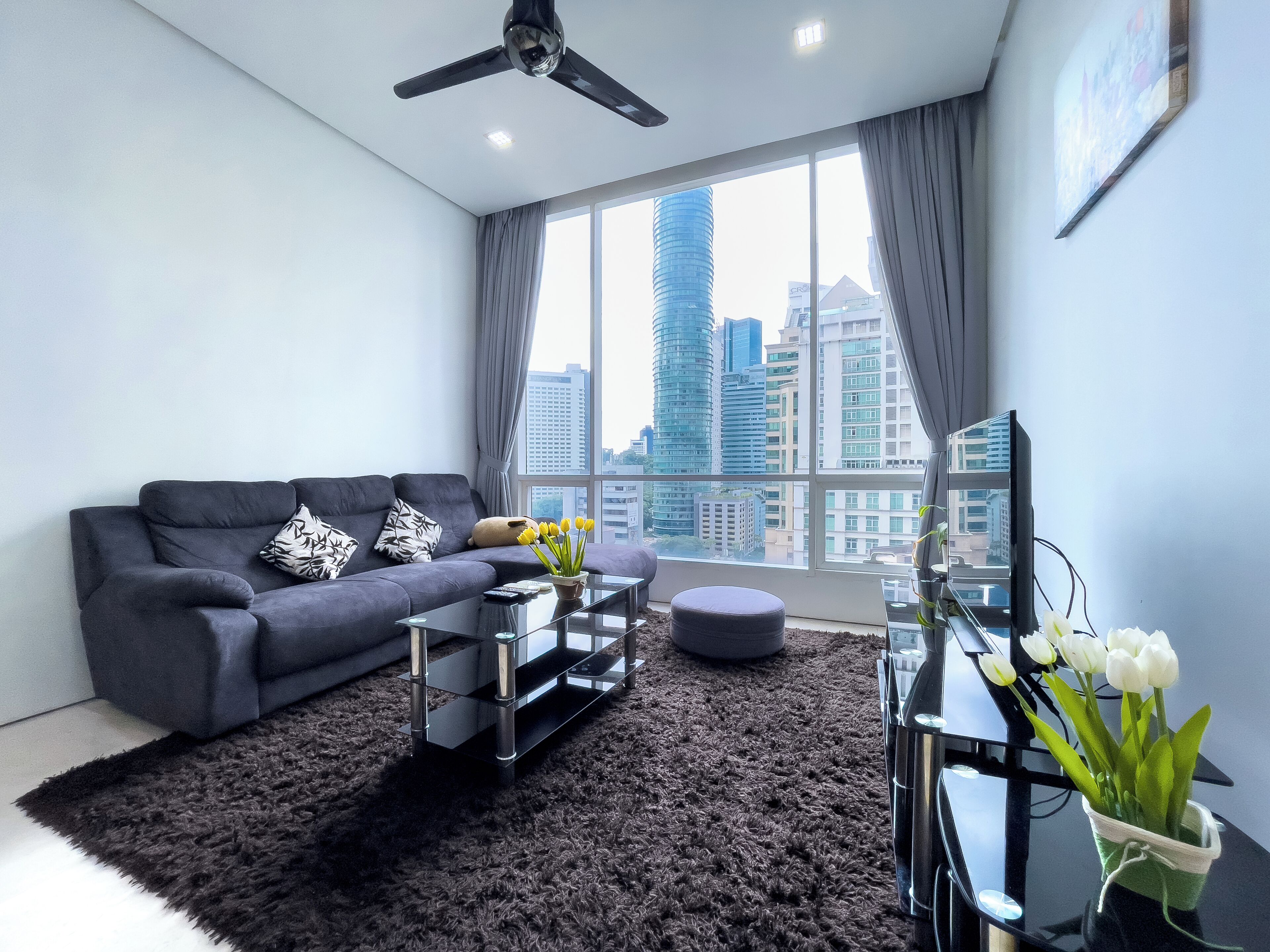 A modern living room with a grey sofa, a glass coffee table, a black ottoman, and a view of city buildings through large windows.