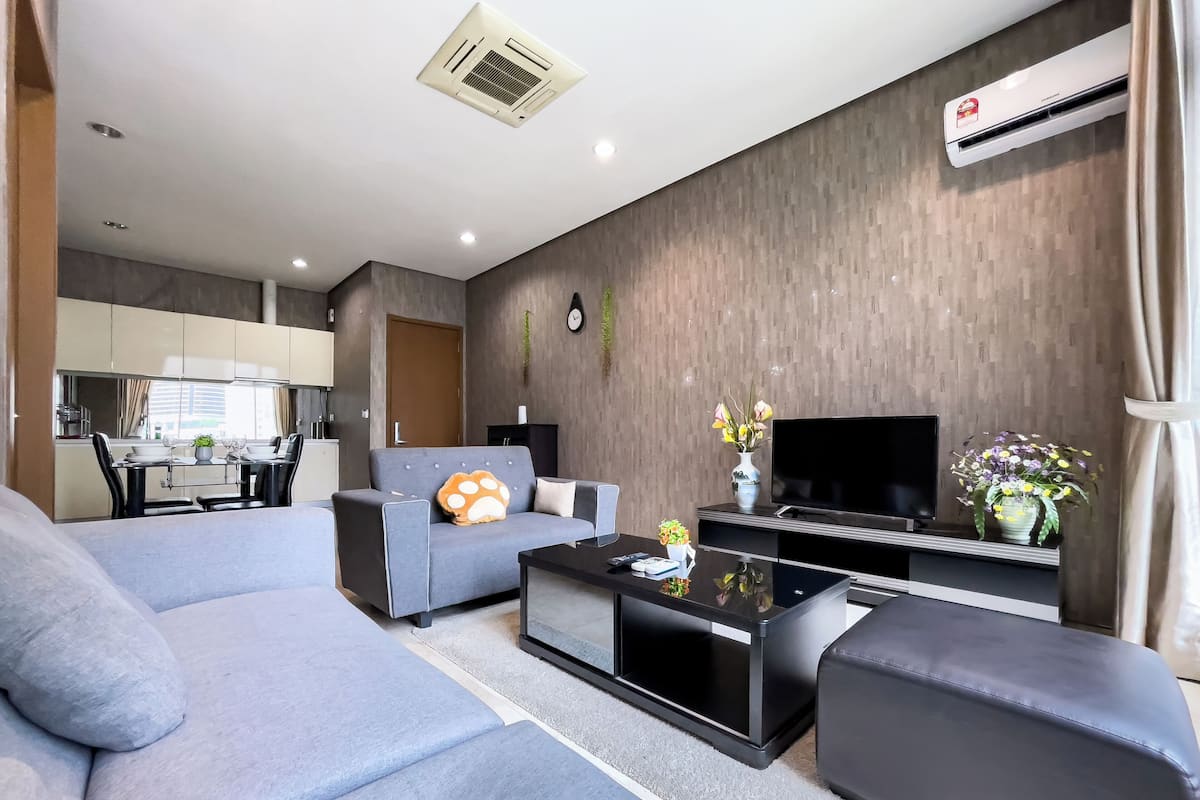 Un salon moderne avec un canapé gris, une table basse noire et un téléviseur à écran plat. On aperçoit une cuisine à l’arrière-plan, avec un coin repas.