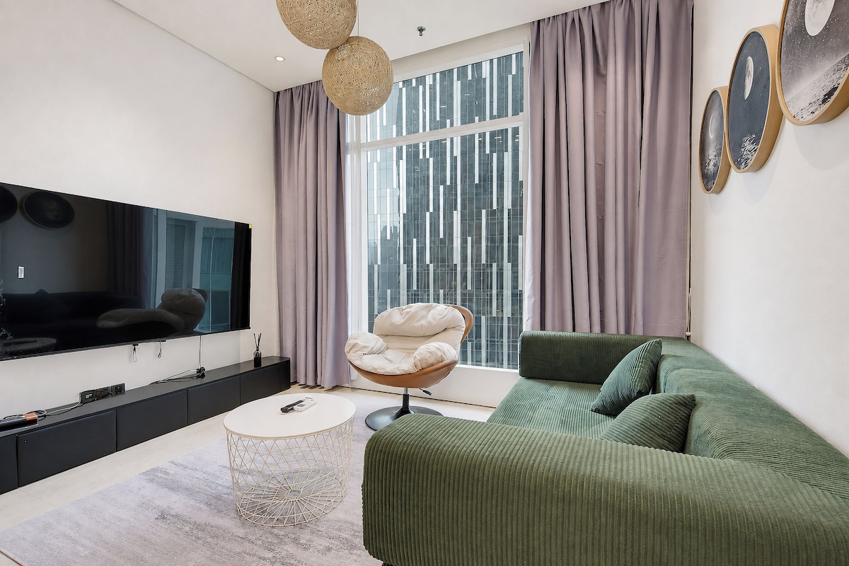 Een moderne woonkamer met een flatscreen televisie, een ronde salontafel, een groene bank en een groot raam met gordijnen.