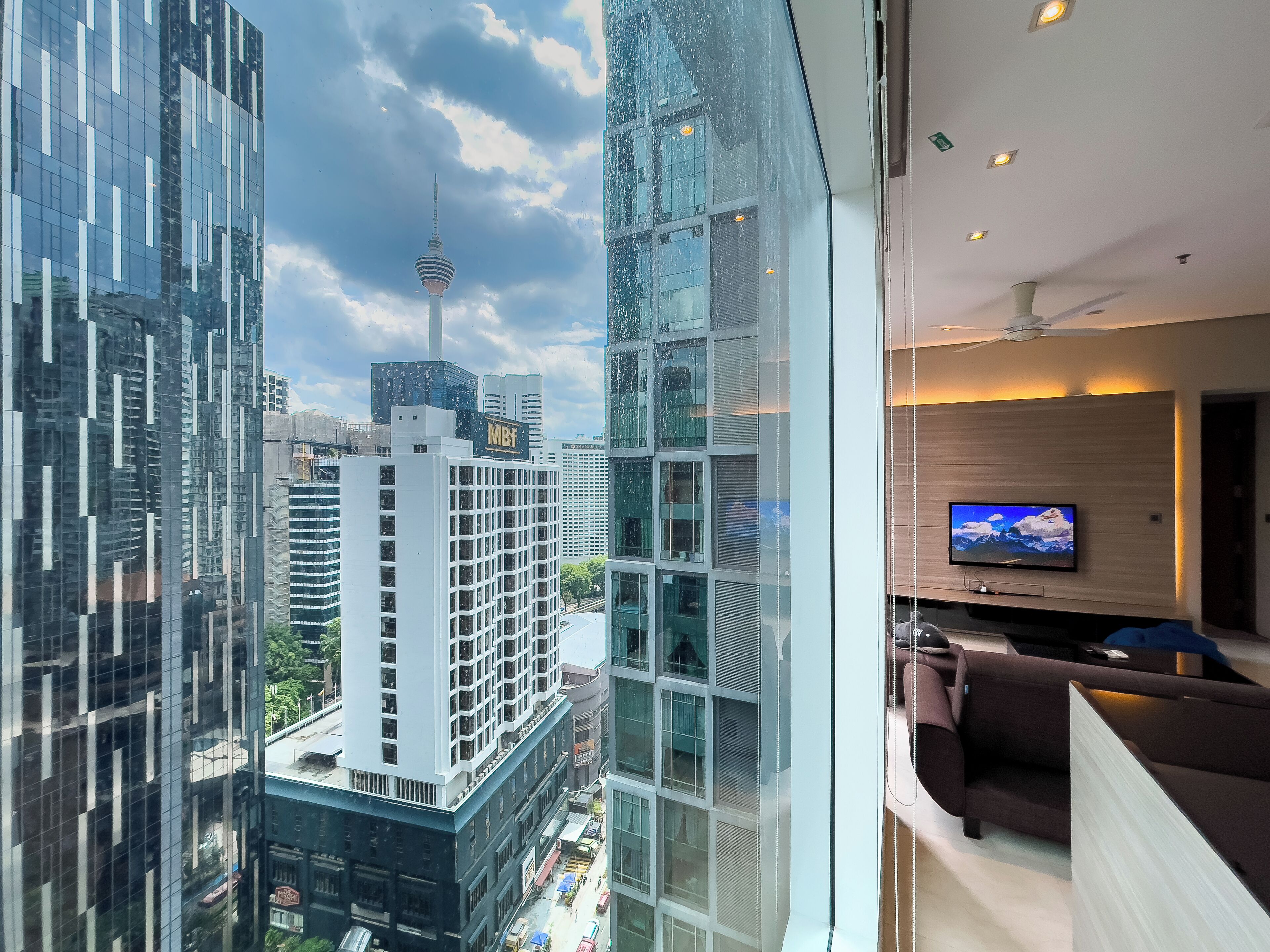 Een moderne hotelkamer met een groot raam dat uitzicht biedt op de stad, een flatscreen televisie aan de muur en een gezellige zithoek met een bank en fauteuils.