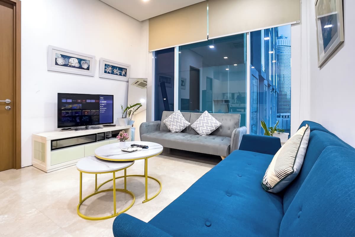 Un salon moderne avec un canapé gris, un canapé bleu, une petite table blanche et un téléviseur posé sur un meuble blanc.