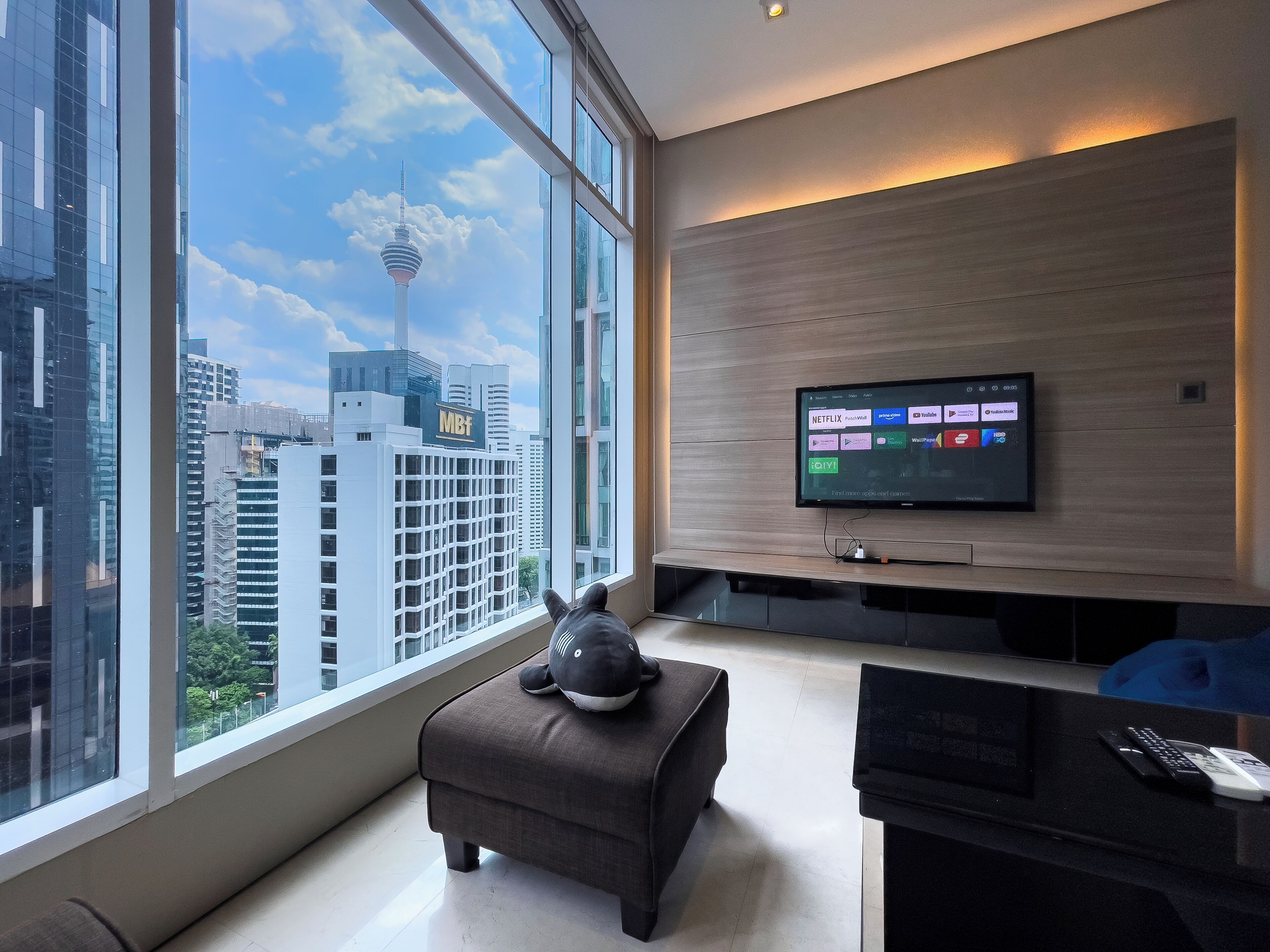 Une chambre d’hôtel moderne dotée d’une grande fenêtre donnant sur la ville, d’un téléviseur à écran plat fixé au mur et d’un confortable pouf.