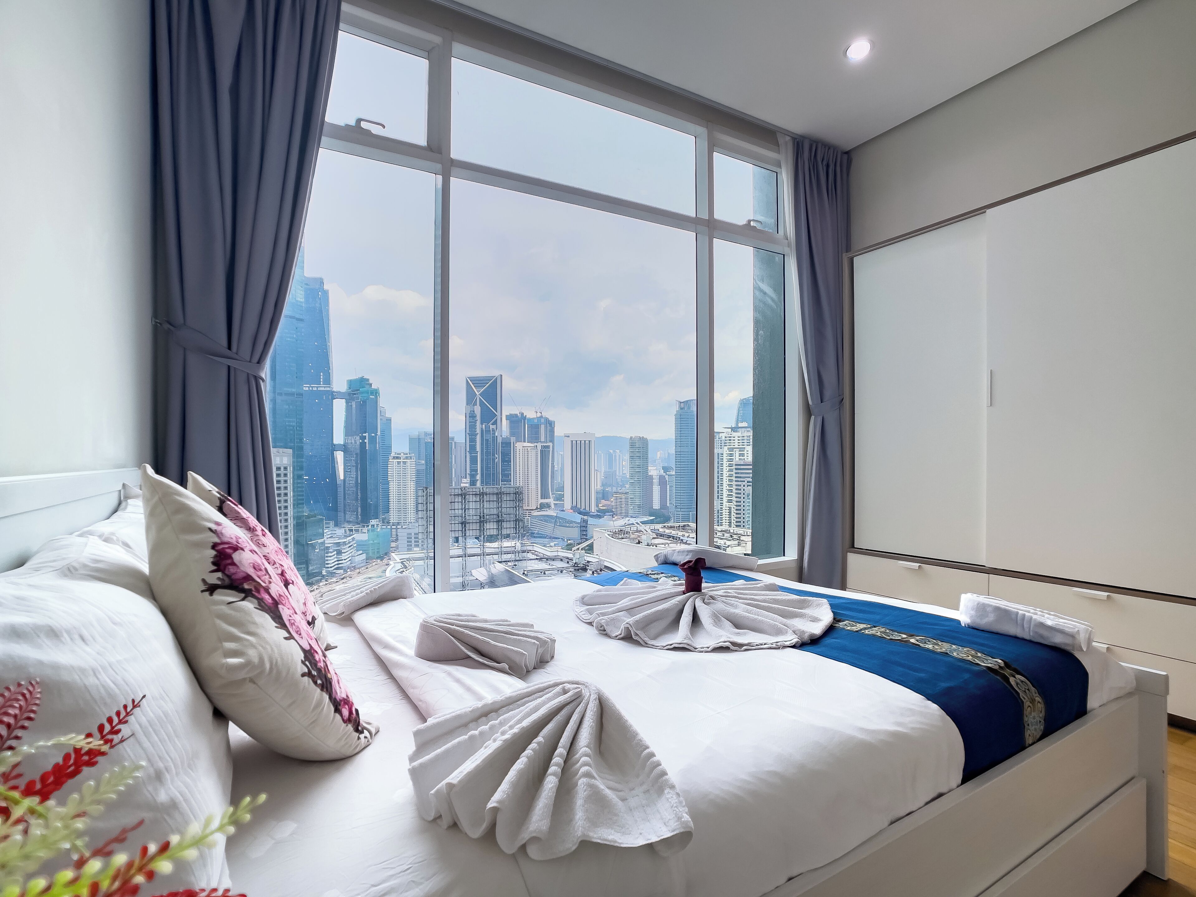 Een hotelkamer met een groot raam met uitzicht op de stad, een net opgemaakt bed met witte lakens en een blauw accent op het beddegoed.