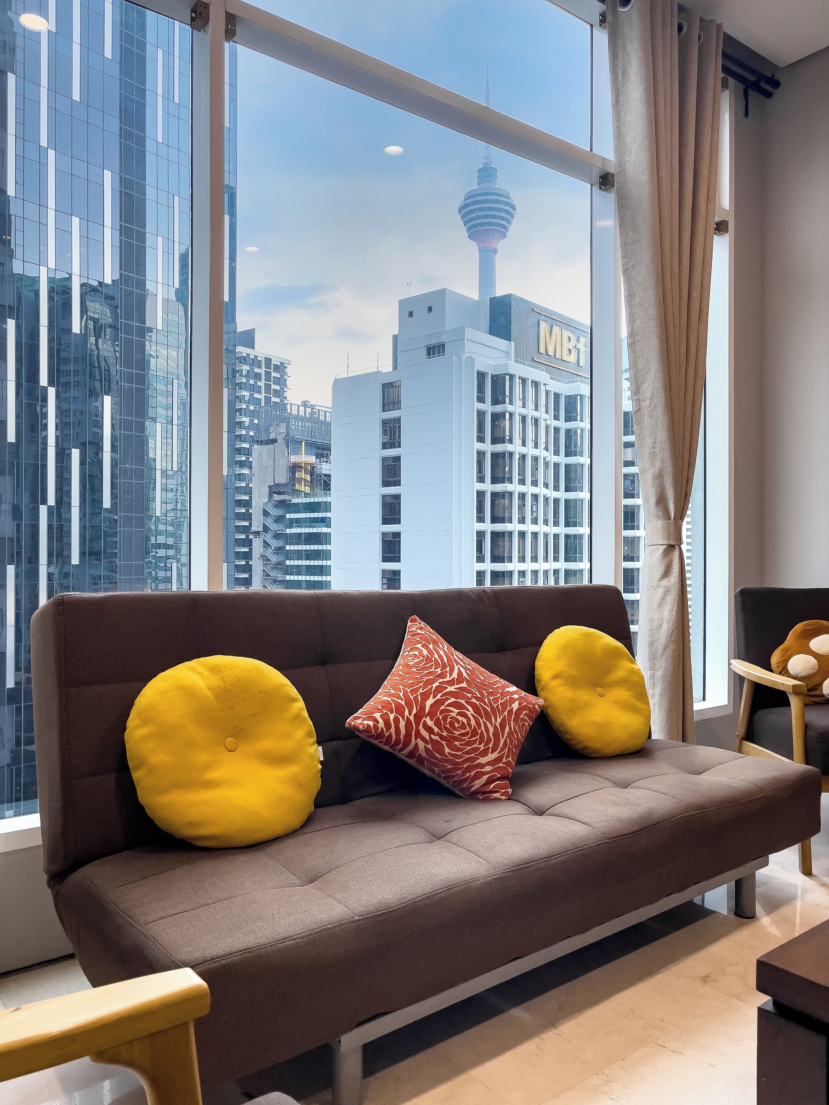 A modern living room with a brown sofa, yellow and red patterned cushions, and a view of a cityscape including a tower and buildings.
