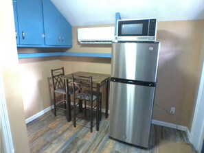 Cocina compacta con refrigerador de acero inoxidable, microondas y una mesa de comedor con cuatro sillas. Los armarios son azules sobre la encimera, y el piso tiene un diseño que imita la madera.