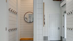 Un couloir aux murs recouverts de panneaux blancs, un banc en bois, un miroir rond, une porte blanche et un paillasson noir.