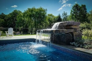 A swimming pool with a waterfall feature, surrounded by greenery and a white chair. - Room with private bathroom (Bromont)
