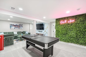 Ein Zimmer mit Tischtennisplatte, grünem Sofa, Flachbildfernseher und einer grünen Wand mit einer Neonreklame.