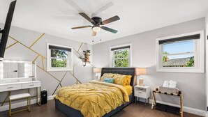 A bedroom with a bed, desk, ceiling fan, and windows.