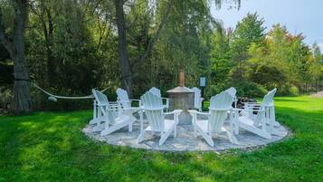 Un espace extérieur circulaire aménagé avec des chaises Adirondack blanches et un foyer central, le tout entouré d’une terrasse en pierre et d’une végétation luxuriante.