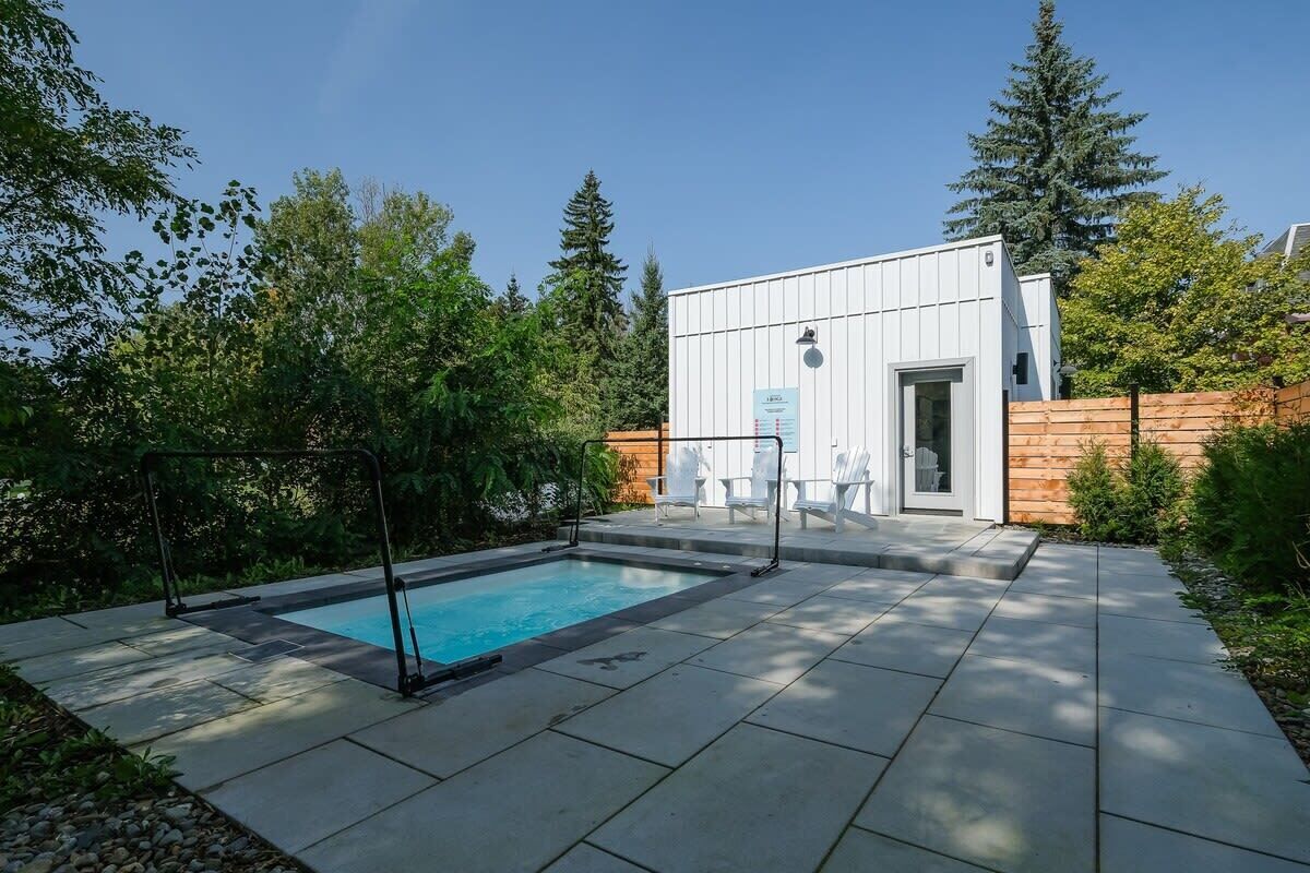 Un espace piscine extérieur moderne, doté d’une terrasse en bois, d’un bâtiment blanc avec une porte vitrée et entouré de verdure.