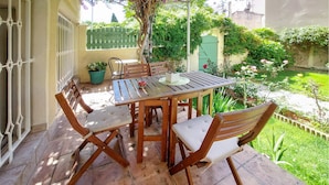 Un patio al aire libre con una mesa y sillas de madera, rodeado de follaje y una valla verde.